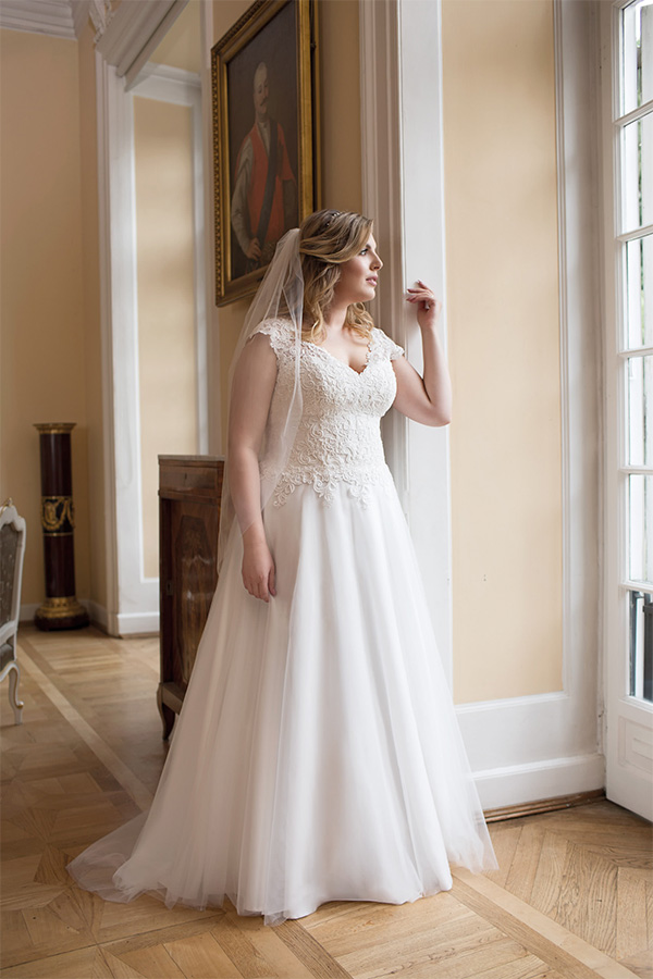 Eine blonde Frau im weißen Brautkleid blickt nachdenklich aus einem Fenster