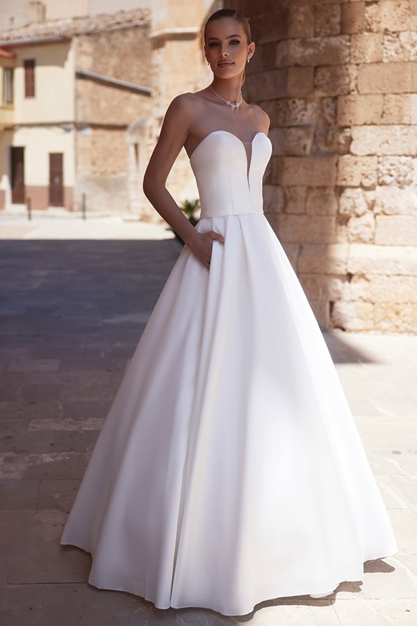 Eine blonde Frau mit festlicher Miene trägt ein elegantes weißes Brautkleid draußen