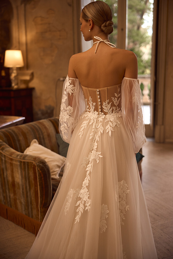 Eine elegant gekleidete Frau steht mit dem Rücken im weißen Brautkleid mit Spitzenornamenten