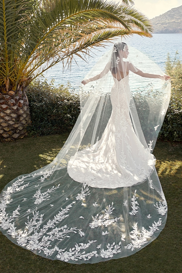 Eine Frau im weißen Brautkleid mit langem Schleier steht lächelnd am See im Grünen