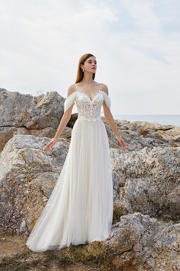 Eine junge Frau mit ernstem Gesichtsausdruck trägt ein elfenbeinfarbenes Spitzenkleid vor Felsen am Meer