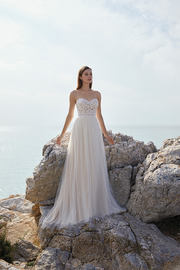 Eine junge Frau in weißem Brautkleid steht entspannt auf Felsen am Meer im Sonnenlicht