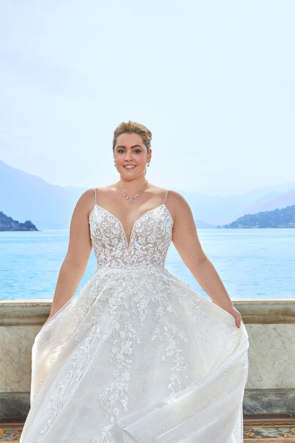 Eine lächelnde Frau im weißen Brautkleid steht vor einem See mit Bergen im Hintergrund