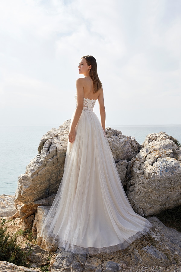 Junge Frau im cremefarbenen Brautkleid lächelt entspannt auf Felsen am Meer