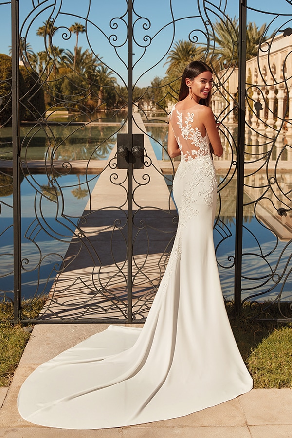 Eine brünette Frau im eleganten weißen Spitzenkleid lächelt vor einem dekorativen Tor