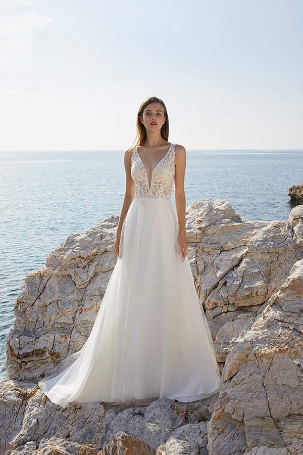 Eine junge Frau mit neutralem Gesichtsausdruck steht in weißem Brautkleid auf Felsen am Meer