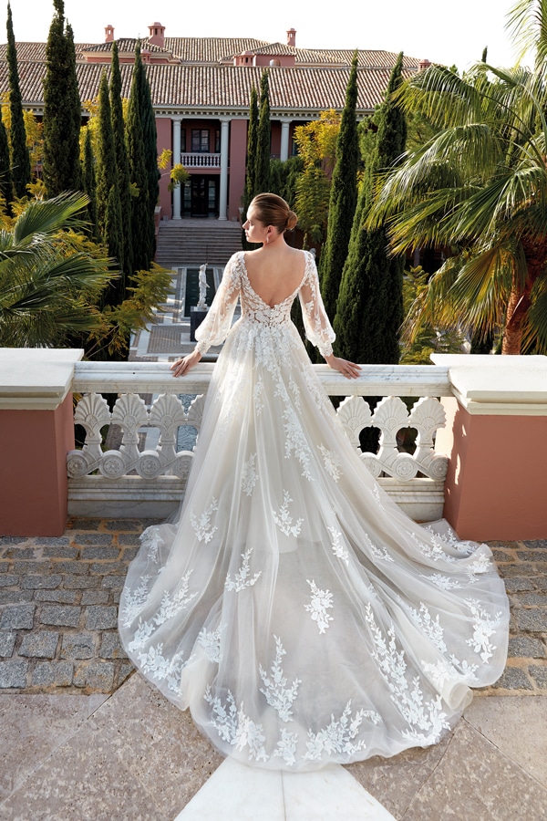 Eine Frau in weißem Spitzenkleid steht auf einem Balkon und blickt auf eine Villa