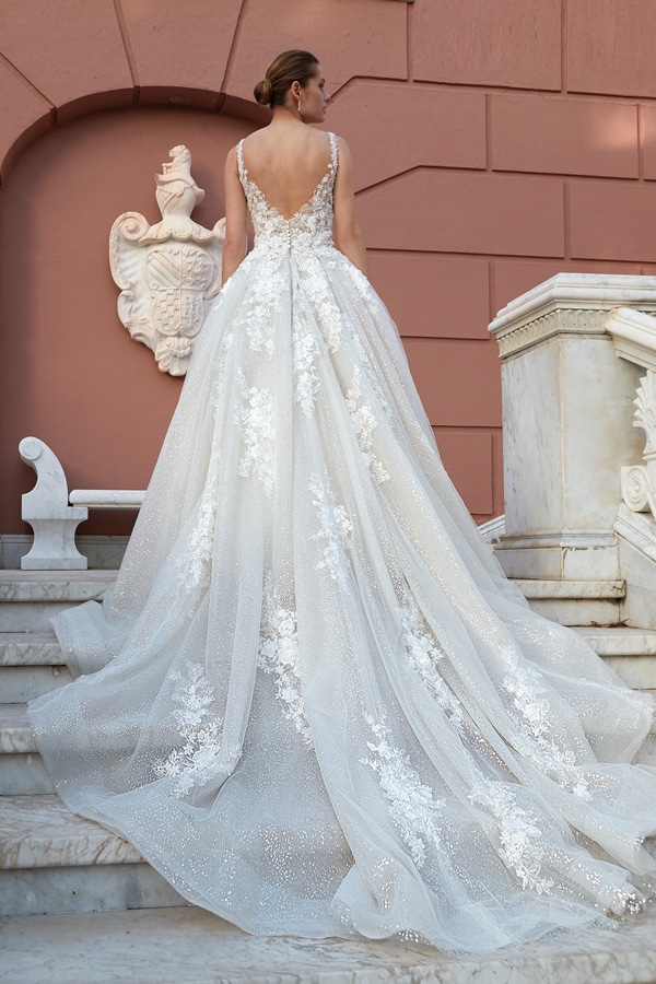 Eine elegante Frau mit Hochsteckfrisur trägt ein funkelndes Brautkleid mit Schleppe und Spitzenapplikationen auf einer Marmortreppe vor einer rosafarbenen Wand