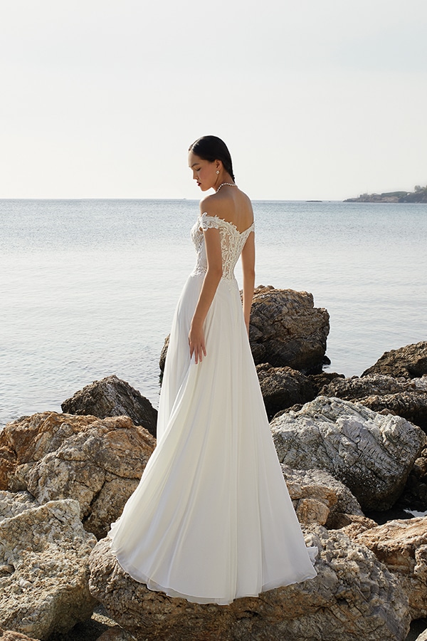 Eine Frau im eleganten weißen Brautkleid steht ruhig vor felsiger Küste und Meer