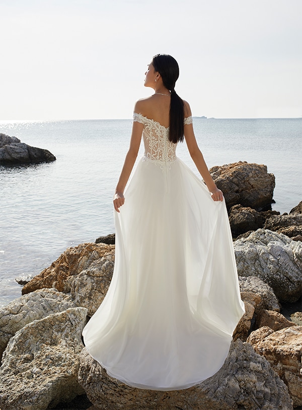 Eine Frau in weißem Spitzenkleid steht mit gelassenem Blick am felsigen Meeresufer