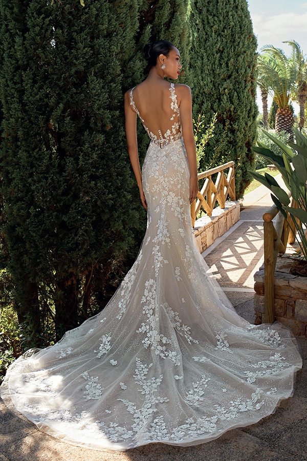 Eine Frau im eleganten, bestickten weißen Brautkleid steht vor grünen Bäumen