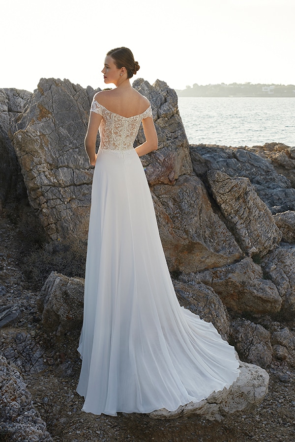 Eine Frau mit strengem Dutt trägt ein elegantes weißes Spitzenkleid am Felsstrand