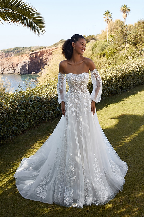 Eine lächelnde Frau in einem weißen Spitzenkleid steht im Sonnenschein im Garten