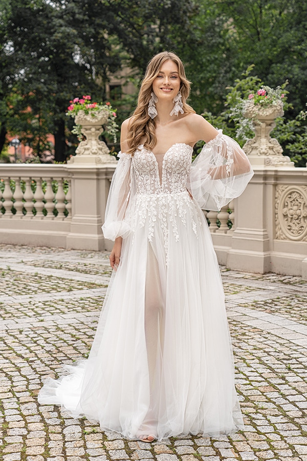 Junge Frau mit lockigem Haar lacht in weißem Brautkleid mit Spitzenärmeln im Park