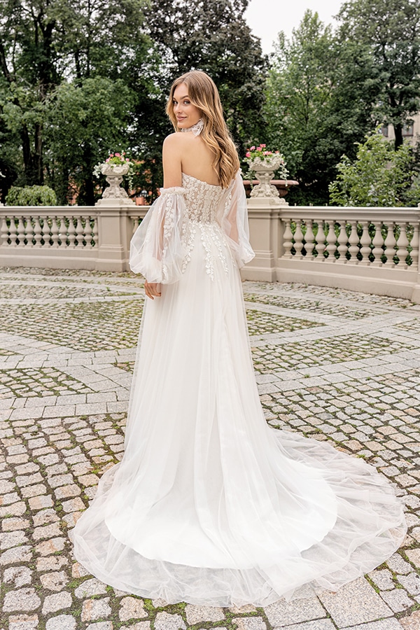 Eine lächelnde Frau im cremefarbenen Brautkleid mit Spitzenärmeln und Schleppe draußen