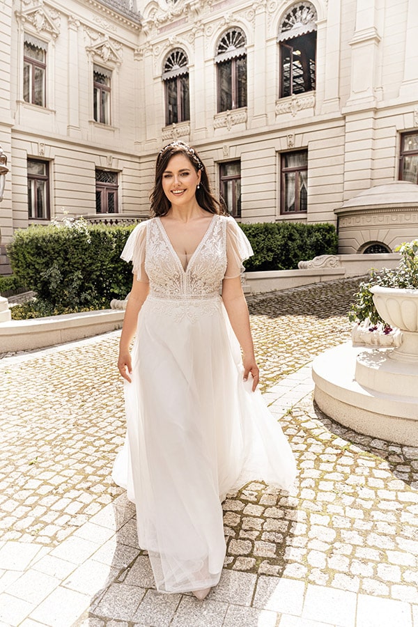 Eine lächelnde Frau im weißen Brautkleid geht im Sonnenschein vor einem historischen Gebäude