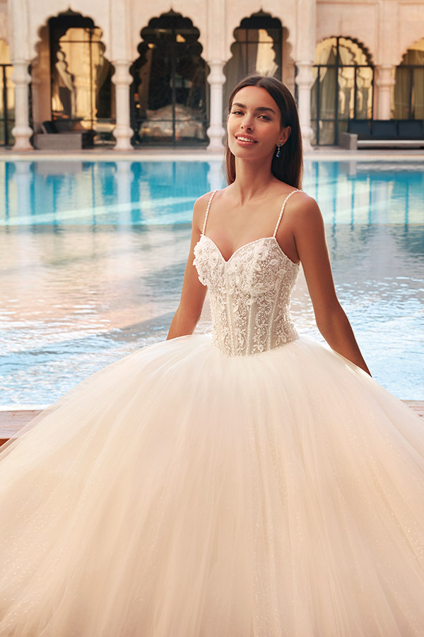 Eine junge Frau mit langem dunklem Haar lächelt sanft in einem weißen Brautkleid vor einem Pool