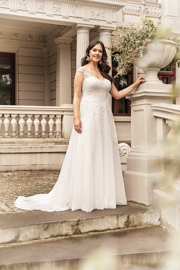 Junge Frau im weißen Brautkleid mit Spitzen, lächelt sanft auf Steintreppe vor Villa