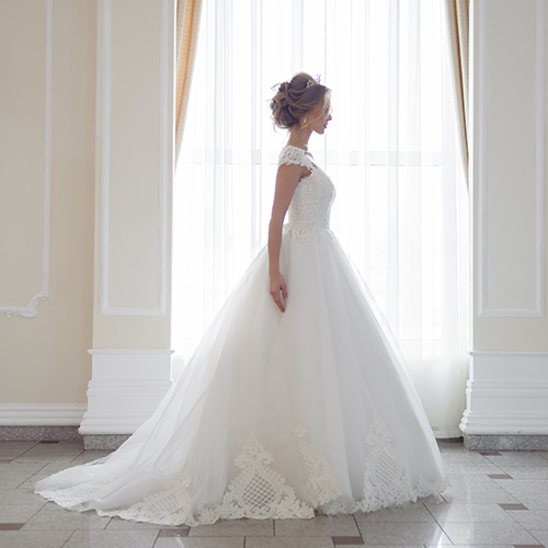 Eine Frau im weißen Brautkleid steht elegant vor einem hellen Fenster
