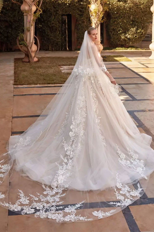 Eine blonde Frau im weißen Brautkleid mit langen Spitzenärmeln schaut ernst zurück