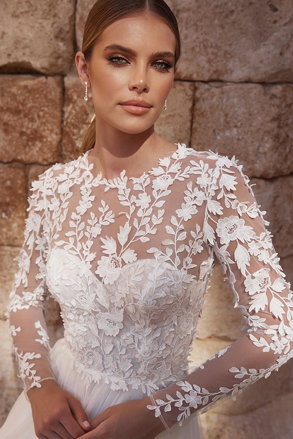 Junge Frau mit neutralem Gesichtsausdruck trägt weißes, florales Spitzenkleid vor Steinwand