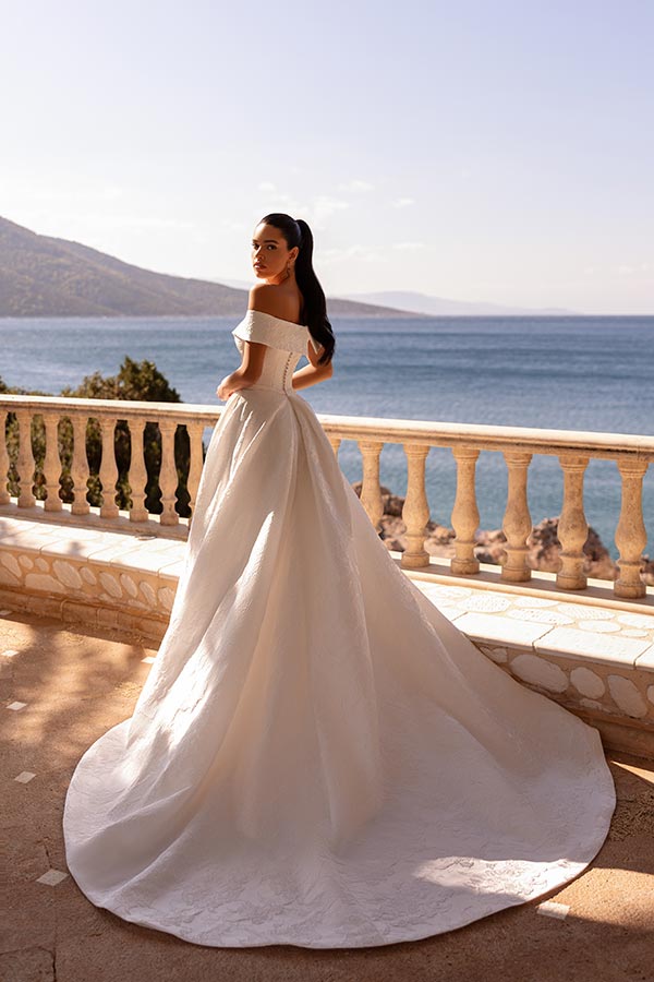Eine Frau in elegantem weißen Brautkleid steht lächelnd auf einer Terrasse am Meer