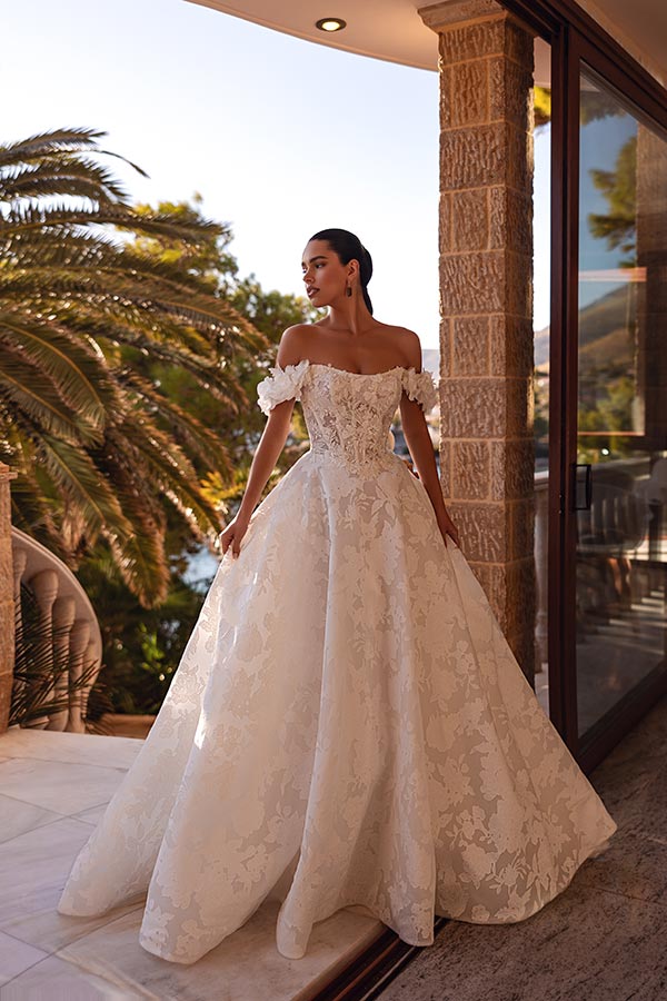 Eine elegante Frau im cremeweißen Brautkleid steht nachdenklich auf einer sonnigen Terrasse