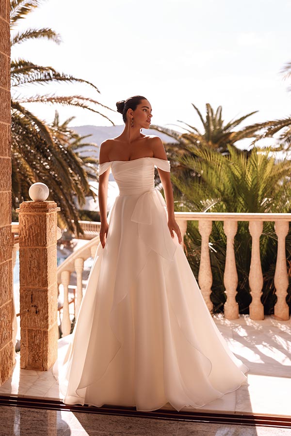 Eine elegant gekleidete Frau im weißen Brautkleid steht nachdenklich auf einer sonnigen Terrasse
