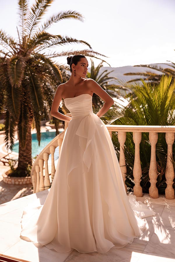 Eine Frau im weißen Brautkleid steht elegant auf einer sonnigen Terrasse mit Palmen