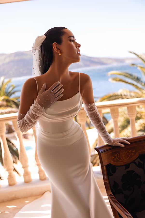 Eine elegant gekleidete Frau im weißen Kleid mit transparenten Handschuhen wirkt nachdenklich auf einem Balkon mit Meeresblick und Palmen im Hintergrund