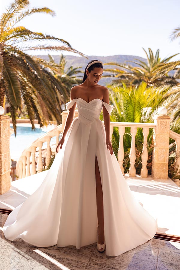 Eine elegante Frau mit ernstem Blick trägt ein weißes, schulterfreies Brautkleid im Sonnenschein