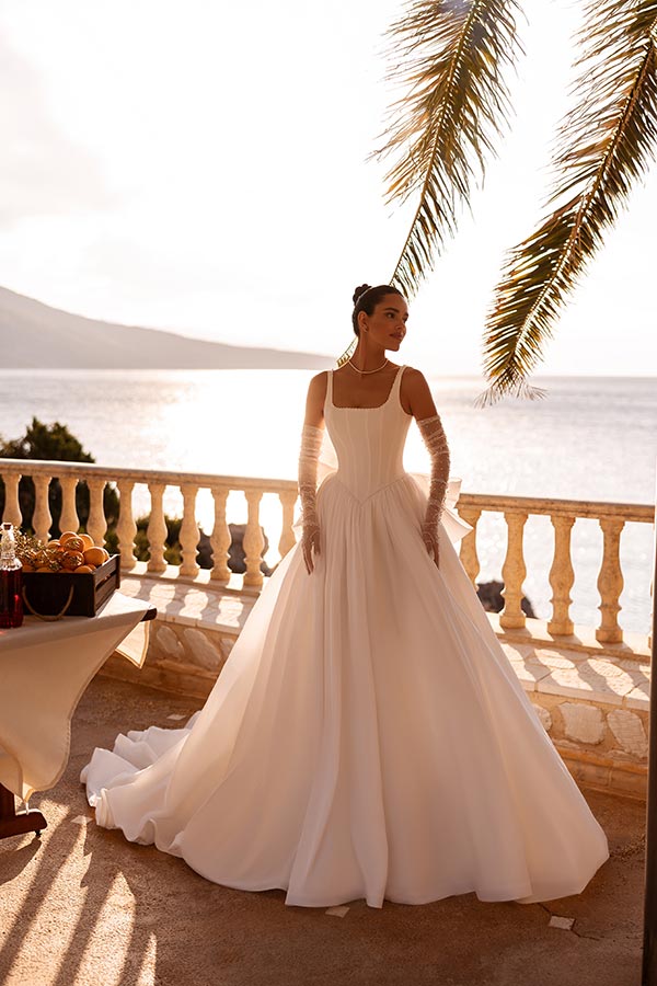 Eine elegante Frau im weissen Brautkleid steht unter einer Palme am sonnigen Meer