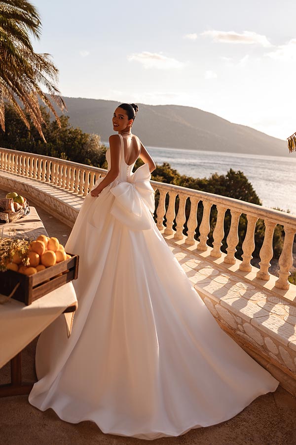 Eine elegante Frau im weißen Brautkleid mit großem Rückenschleifen steht lächelnd auf einer sonnigen Terrasse am Meer