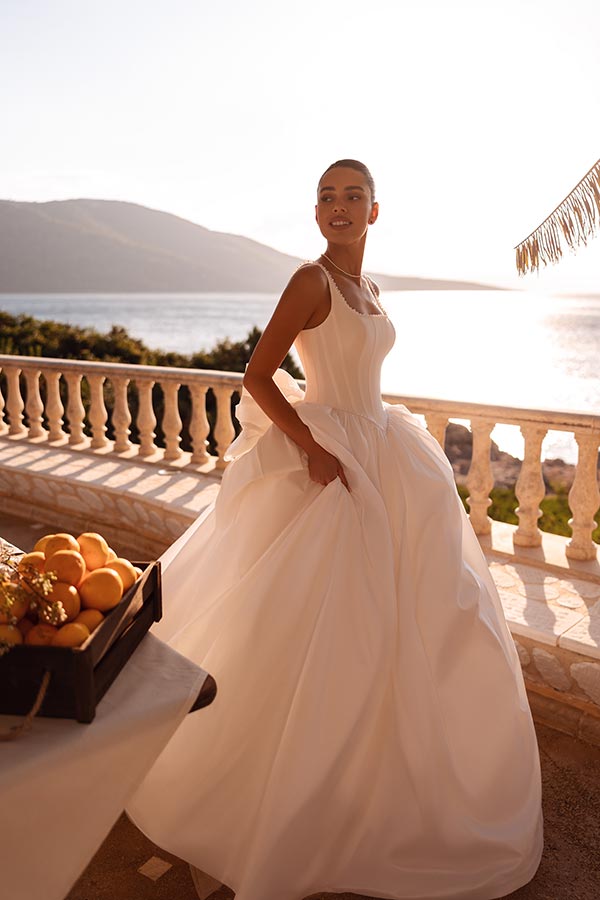 Eine junge Frau mit elegantem Lächeln trägt ein weißes Brautkleid am sonnigen Meer