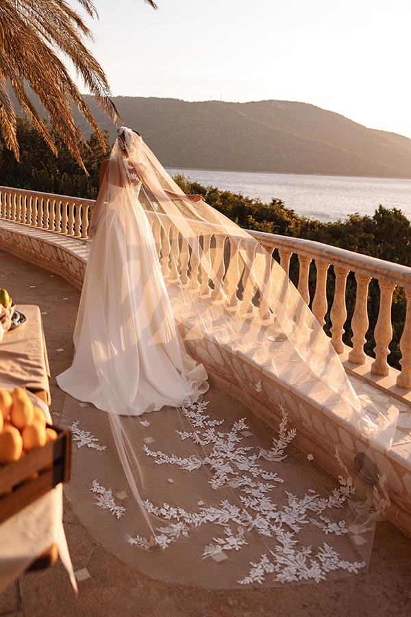 Eine Braut im weißen Kleid mit langem Schleier steht träumend auf einer Terrasse am Wasser