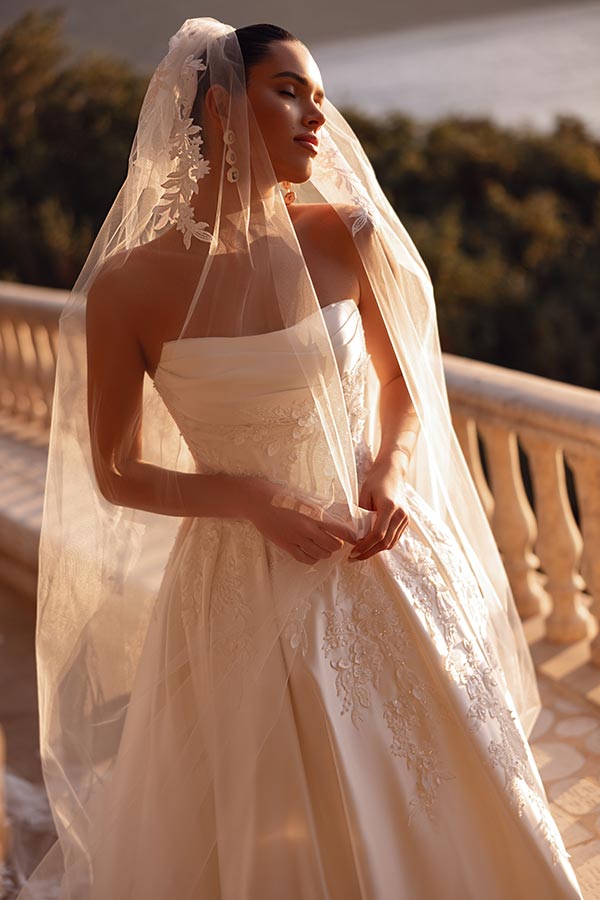Eine lächelnde Frau im eleganten weißen Brautkleid mit Schleier steht draußen am Geländer