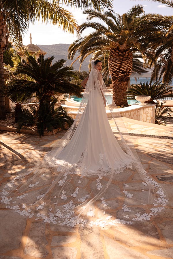 Eine Braut mit ernstem Blick trägt ein elegantes weisses Kleid mit langer Spitze vor Palmen