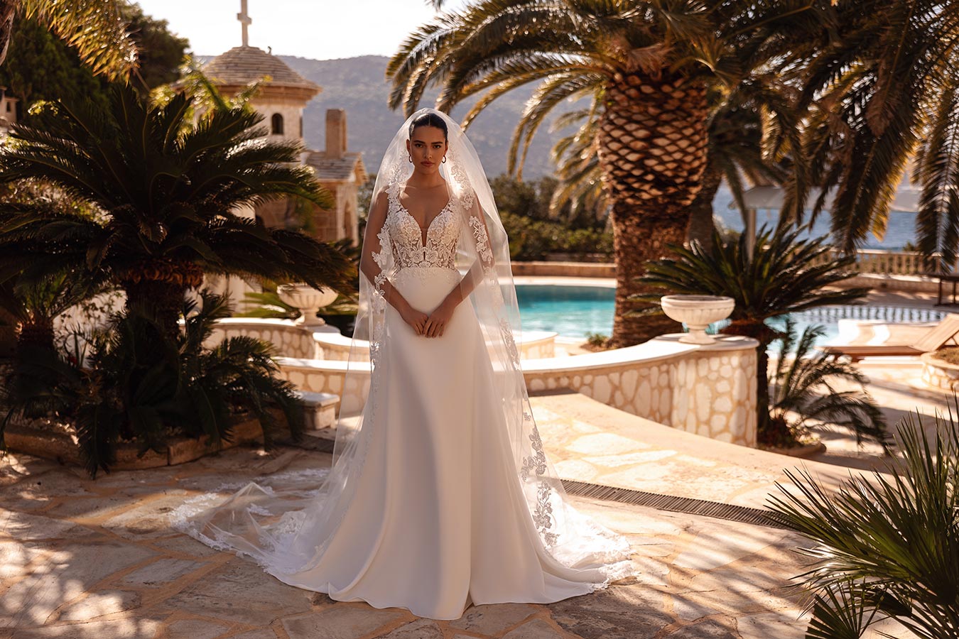 Eine Frau im weißen Brautkleid steht ernst zwischen Palmen vor einem Pool im Sonnenschein