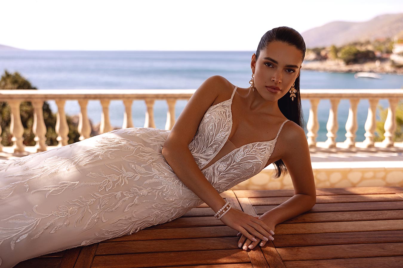 Eine Frau mit ernstem Blick liegt im eleganten weißen Spitzenkleid am Holztisch vor Meereskulisse