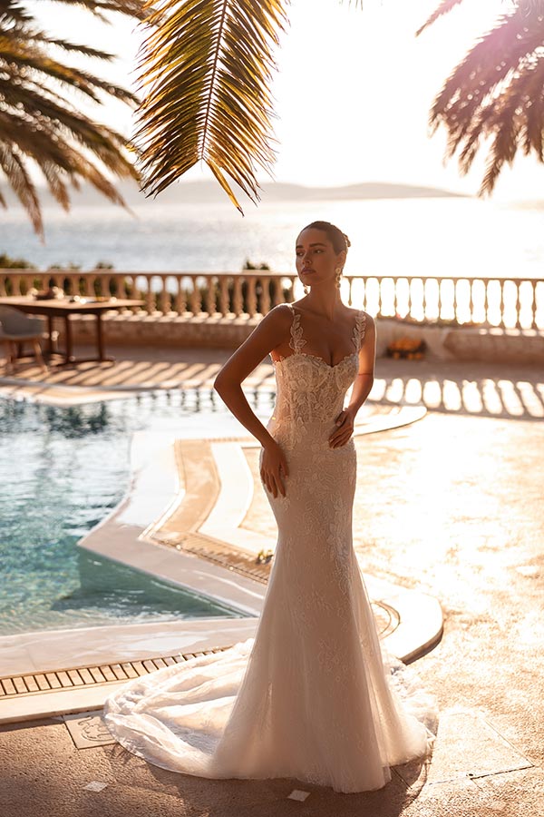 Eine Frau im eleganten cremefarbenen Brautkleid steht nachdenklich am Pool im Sonnenlicht