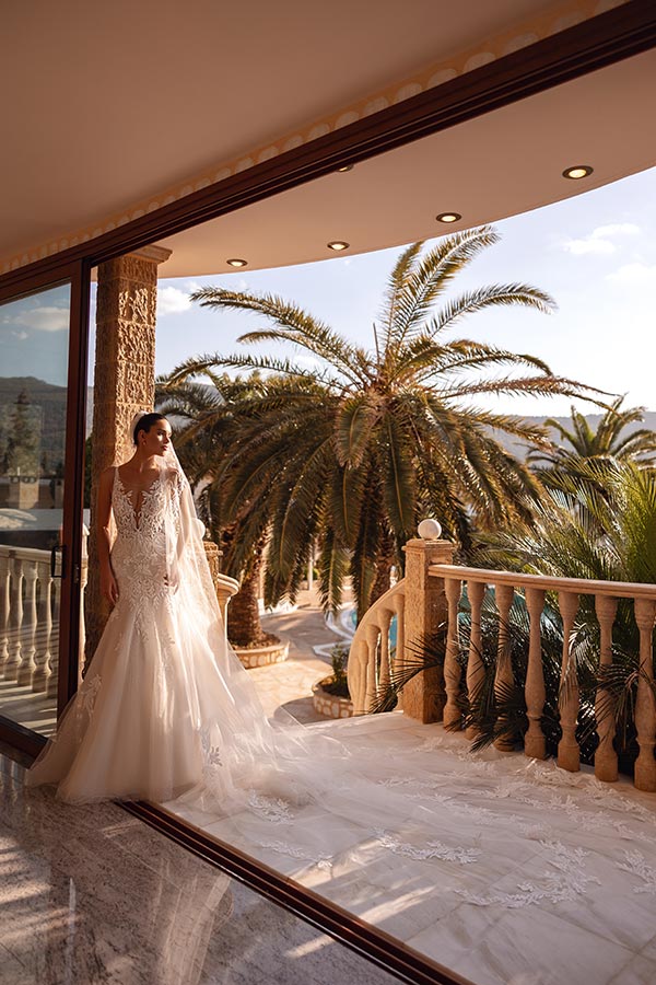 Eine lächelnde Braut im weißen Kleid steht auf einem Balkon mit Palmen im Hintergrund