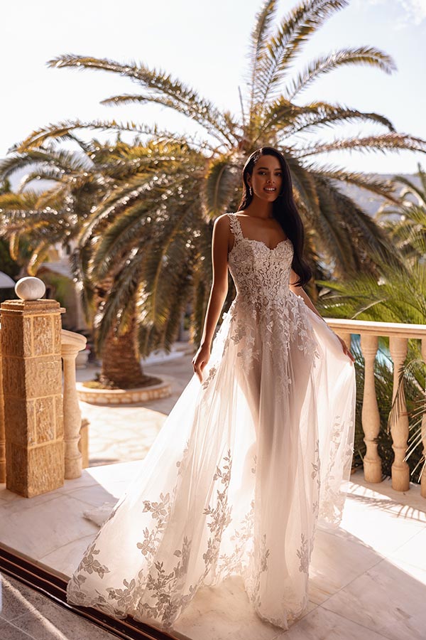 Eine lächelnde Frau in einem eleganten weißen Hochzeitskleid mit Spitzenapplikationen steht vor Palmen im Sonnenschein