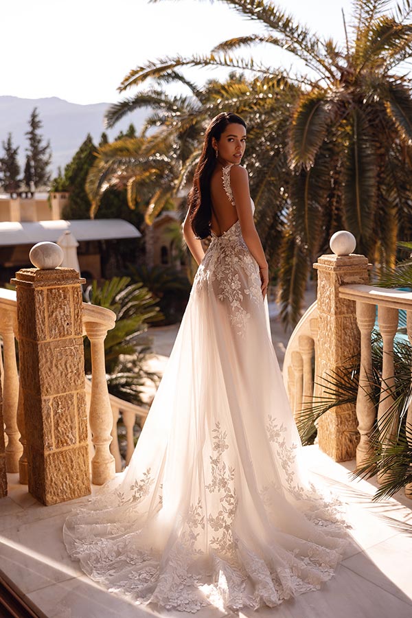 Frau mit ernstem Blick trägt weißes, florales Brautkleid auf sonnigem Balkon