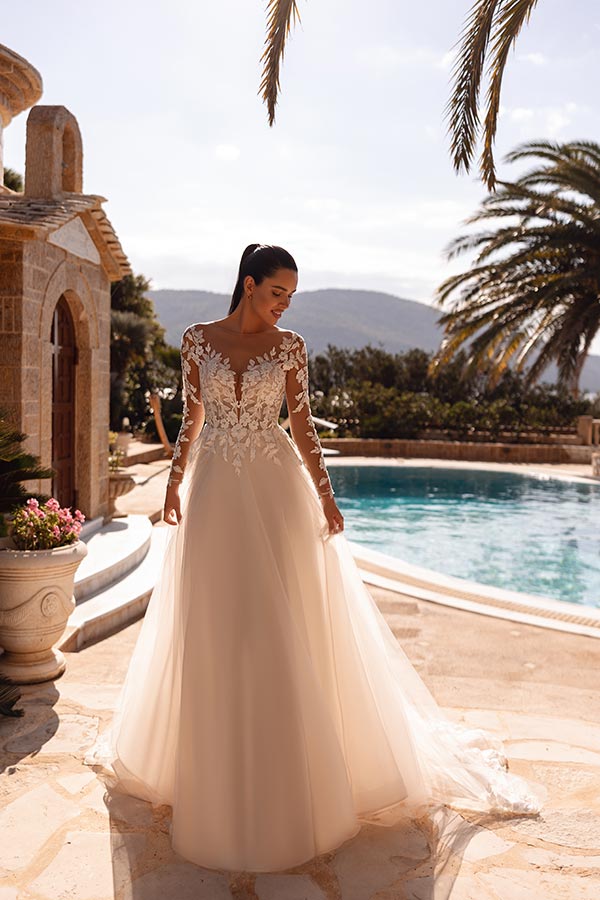 Eine lächelnde Frau im cremefarbenen Spitzenkleid steht elegant am Pool unter Palmen