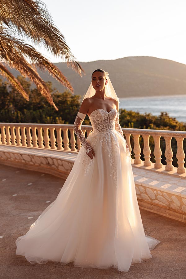 Eine elegant lächelnde Frau im weißen Brautkleid steht auf einer sonnigen Terrasse