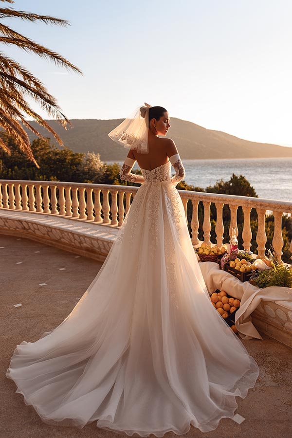 Frau im eleganten weißen Brautkleid steht lächelnd auf Terrasse mit Meerblick