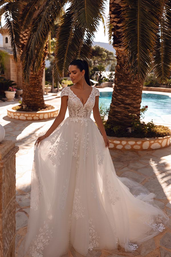 Eine junge Frau mit elegantem Kleid steht lächelnd vor Palmen und Pool im Sonnenlicht
