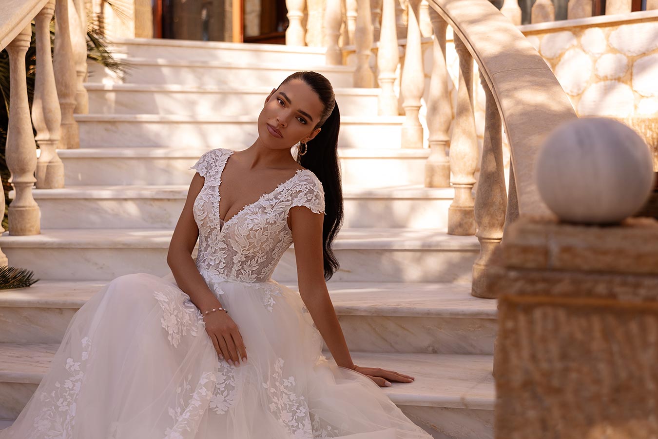 Eine Frau mit langem dunklem Haar und elegantem Brautkleid sitzt lächelnd auf einer Marmortreppe