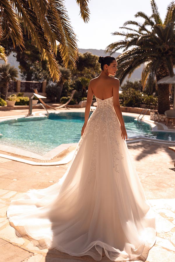 Eine Frau im weißen Brautkleid steht lächelnd an einem sonnigen Pool mit Palmen