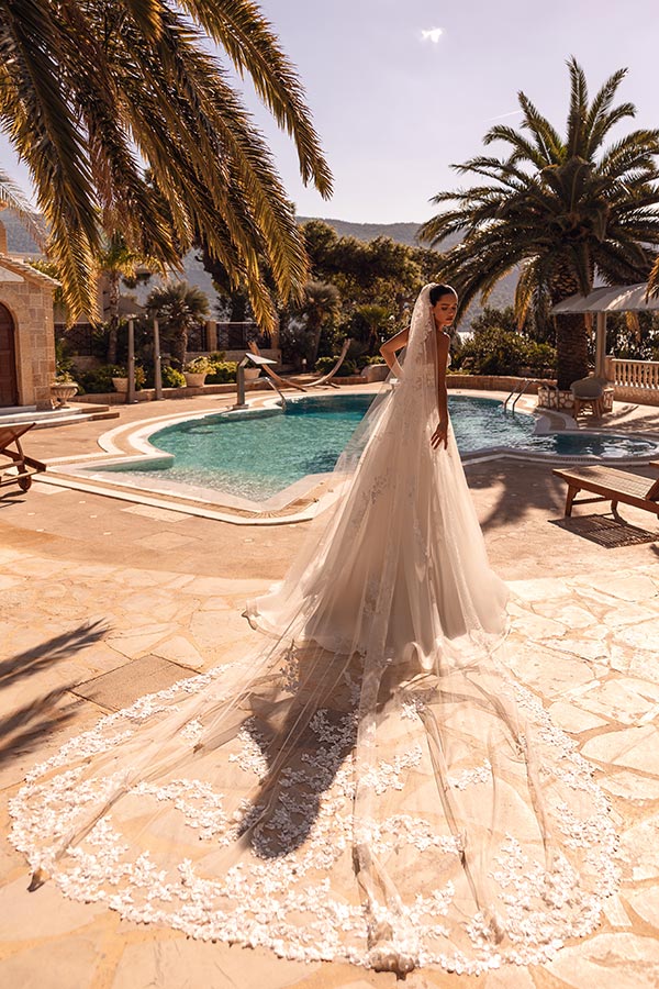 Eine lächelnde Braut mit langem, weißem Spitzenkleid steht am sonnigen Pool